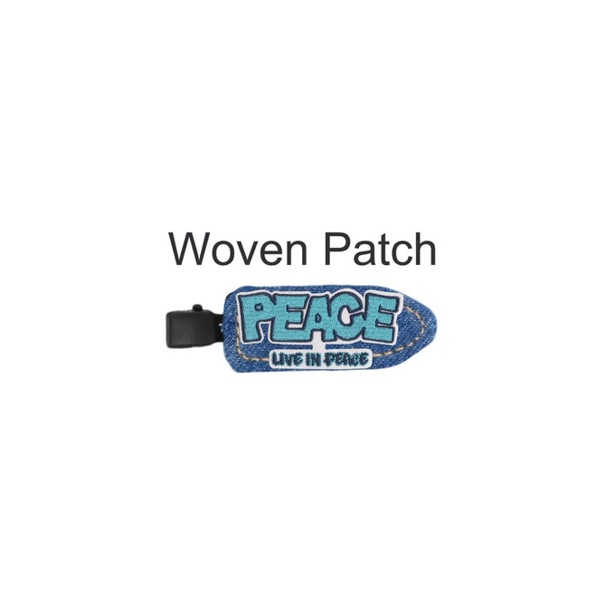 A denim duck bill hair clip in blue with a woven patch displaying the word "PEACE". It features a logo on the clip.