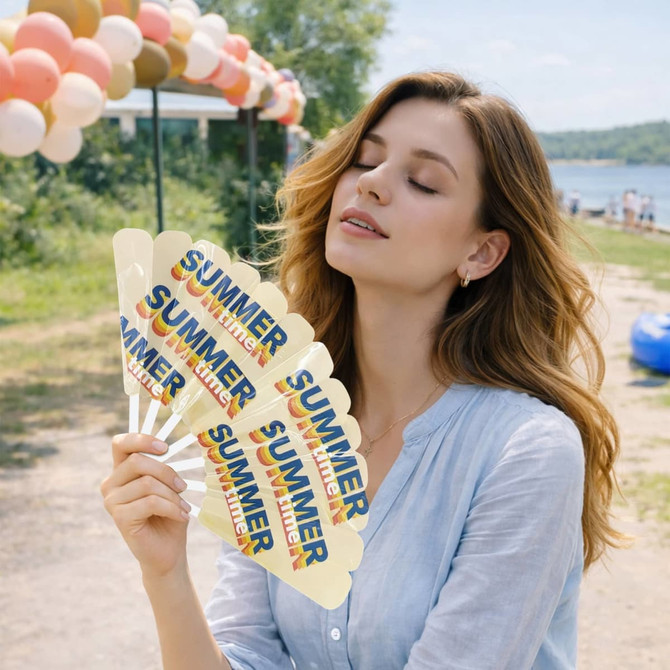 A woman with wavy hair holds multiple yellow plastic hand fans, featuring a colourful design, near a lakeside setting.