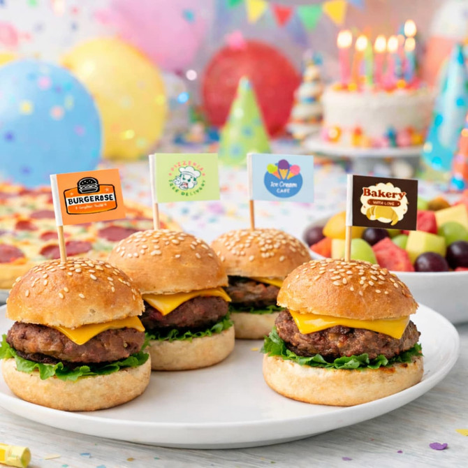 A plate of sesame seed burgers with cheese and lettuce, topped with colourful flags, surrounded by party decorations.