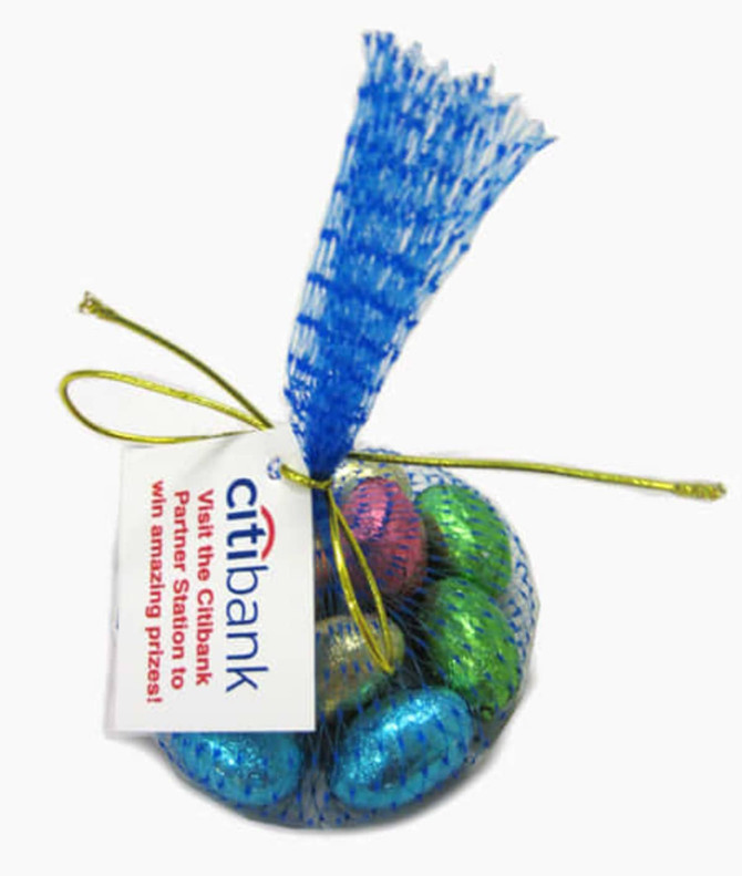 A mesh bag with a blue net contains shiny chocolate Easter eggs in pink, green, and gold. The bag is tied with a gold ribbon.