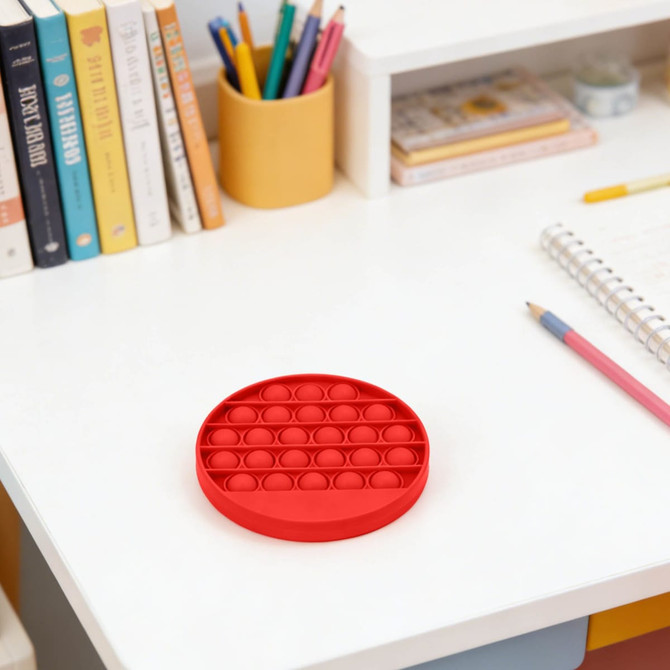 A red round fidget toy with poppable bubbles sits on a white desk beside a notebook and stationery.