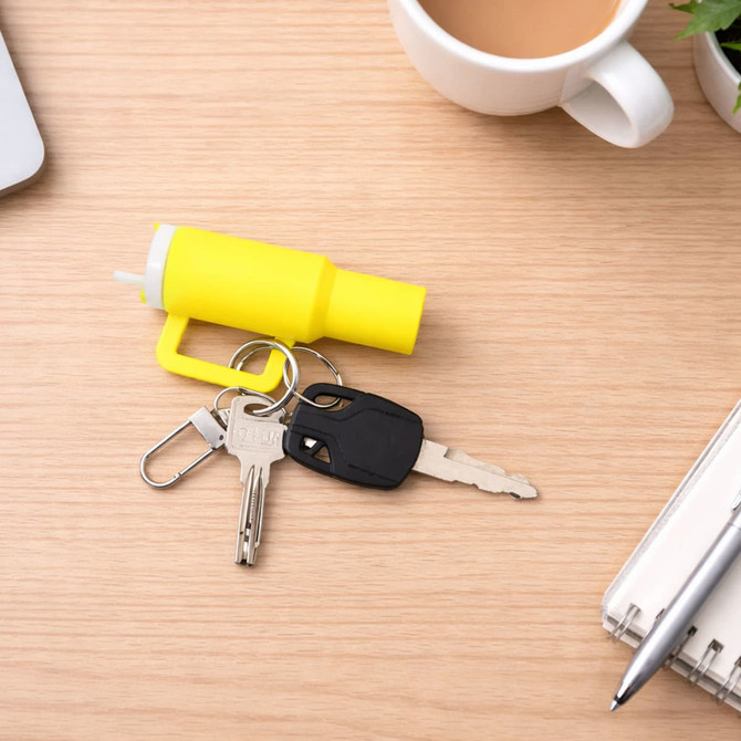 A yellow mini tumbler keyring is placed on a wooden surface next to keys, a pen, and a cup of coffee.