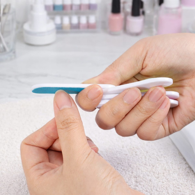 A foldable nail file block in white and blue, held between fingers, with blurred nail polish bottles in the background.