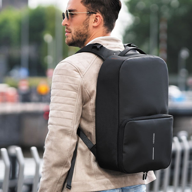 A man wearing a beige jacket carries a sleek black backpack with a logo, set against an outdoor city backdrop.