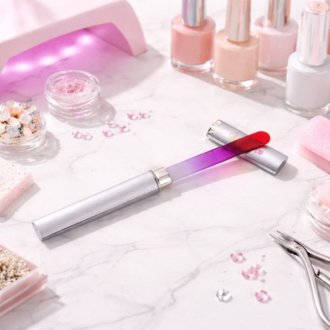 A crystal glass nail file with a silver handle and a gradient pink to red filing surface, surrounded by beauty products.