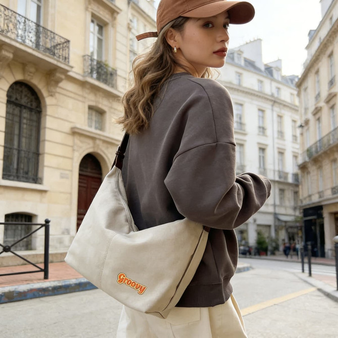 A beige canvas tote bag with a shoulder strap, worn by a person in a grey sweatshirt and tan cap, on a city street.