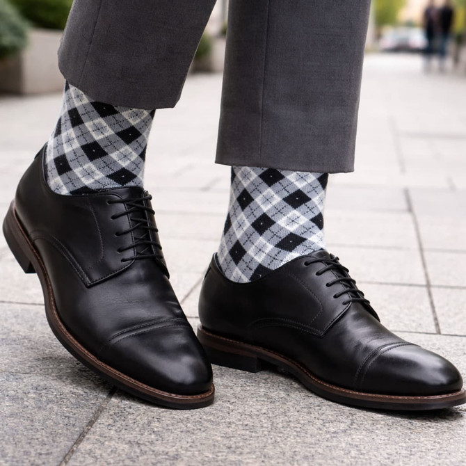 A pair of black leather shoes with grey and black checked socks, set against a stone pavement backdrop.