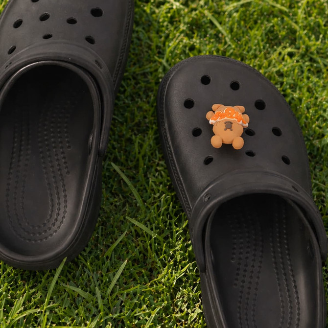A pair of black clog shoes featuring a 3D bear charm on one shoe, set on green grass.