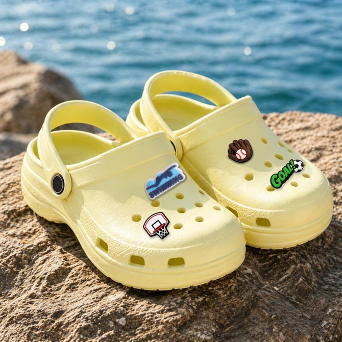 A pair of yellow shoes with various decorative charms, including sports themes, on a rocky surface near water.
