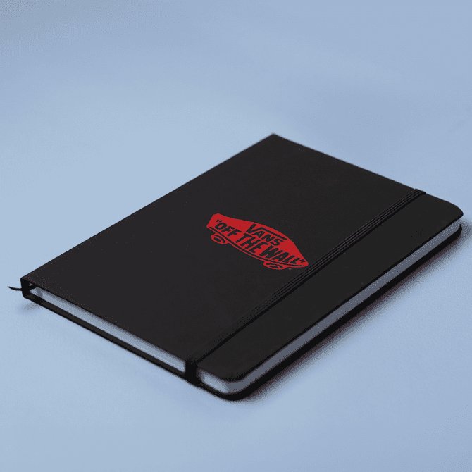 A black A5 notebook with a logo on the cover and an elastic closure, set against a light blue background.