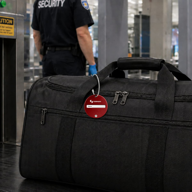 A black luggage bag with a round red wire cable luggage tag attached, placed on a security screening table.