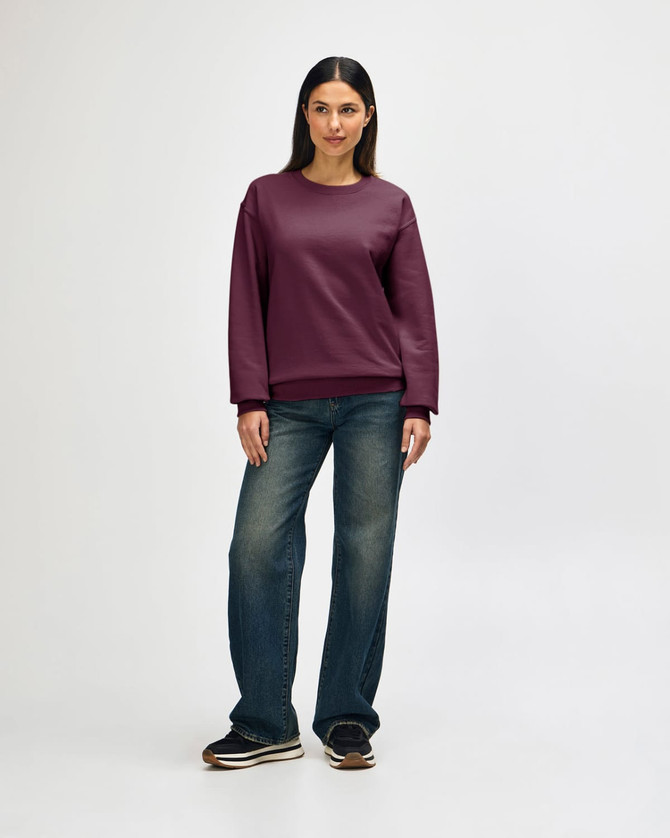 A woman stands wearing a burgundy Gildan Heavy Blend adult crewneck sweatshirt and blue jeans. The sweatshirt features a logo.