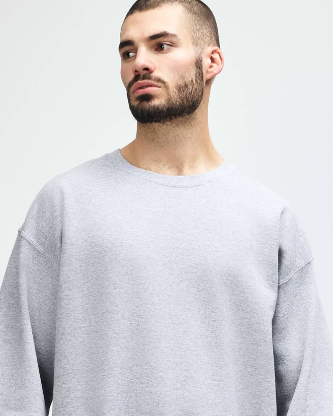A grey Gildan Heavy Blend adult crewneck sweatshirt displayed on a model posing thoughtfully. It features a logo.