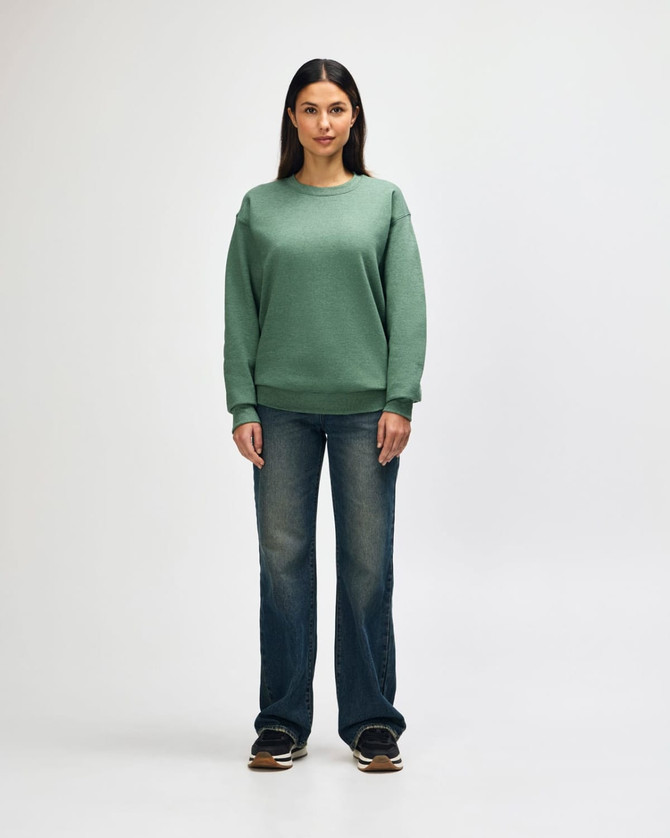 A green Gildan Heavy Blend adult crewneck sweatshirt worn by a model with black jeans and black sneakers. It has a logo.