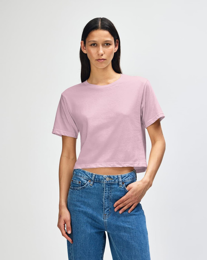 A woman stands wearing a pink boxy t-shirt and blue jeans. The t-shirt has a logo on it.