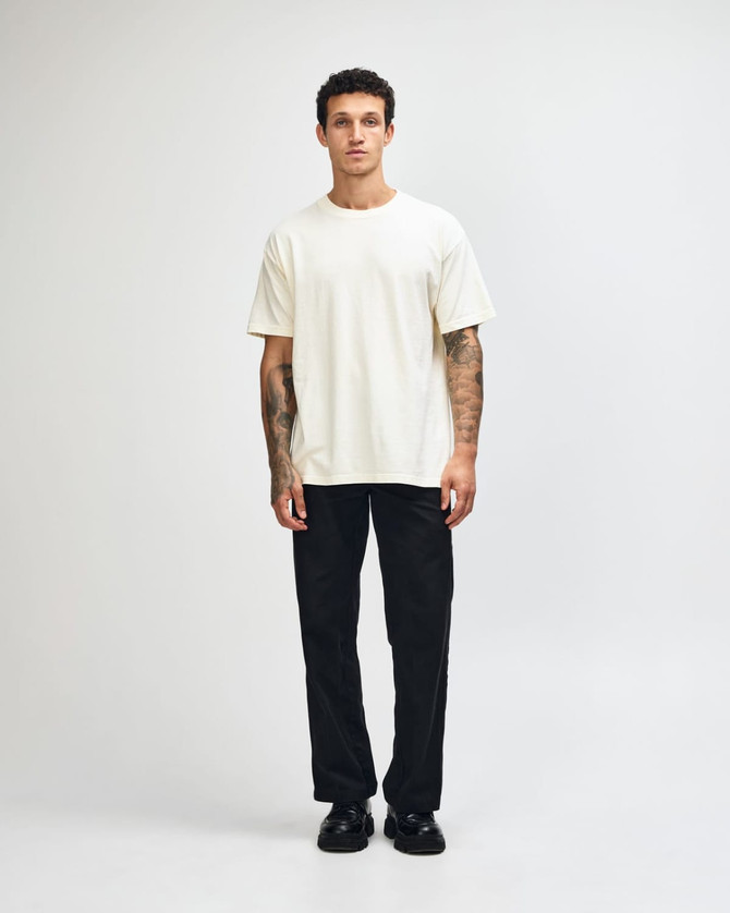 A unisex heavyweight cotton T-shirt in cream, paired with blue cargo pants and sneakers. The model has curly hair and a neutral pose.