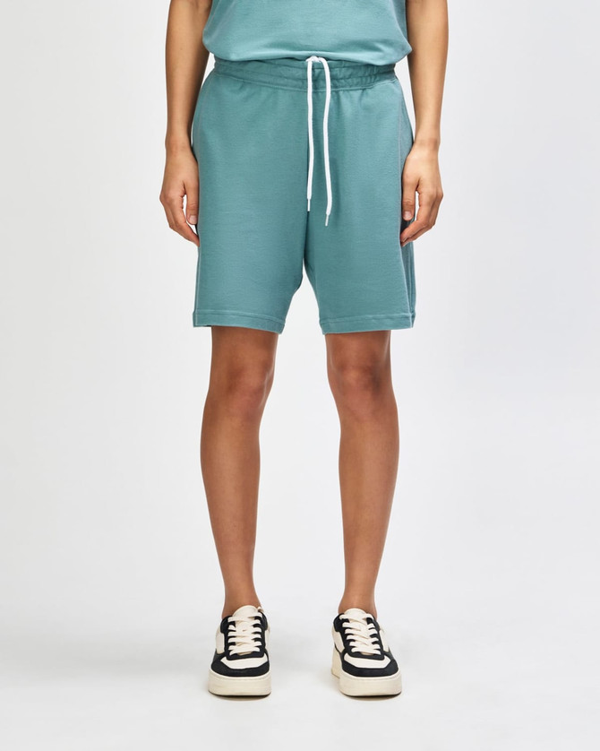 Unisex gym shorts in a soft teal colour, featuring an elastic waistband and pockets, paired with white sneakers.