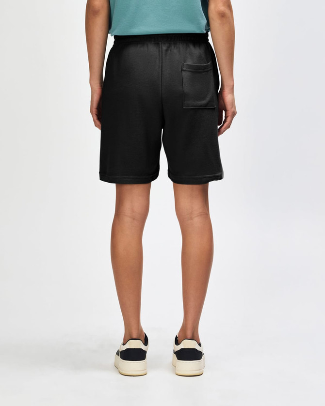 Unisex pique gym shorts in black, featuring an elastic waistband and side pockets, worn with black sneakers and white socks.
