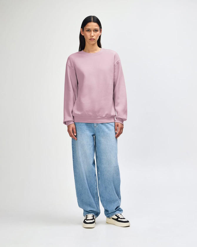A unisex crewneck sweatshirt in light purple, paired with blue jeans and worn by a model. The sweatshirt has a branding logo.