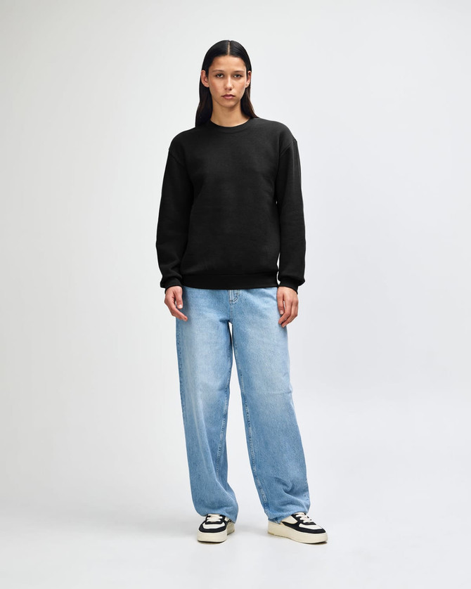 A unisex crewneck sweatshirt in black, paired with light blue cargo pants, featuring curly hair. The sweatshirt has a logo.