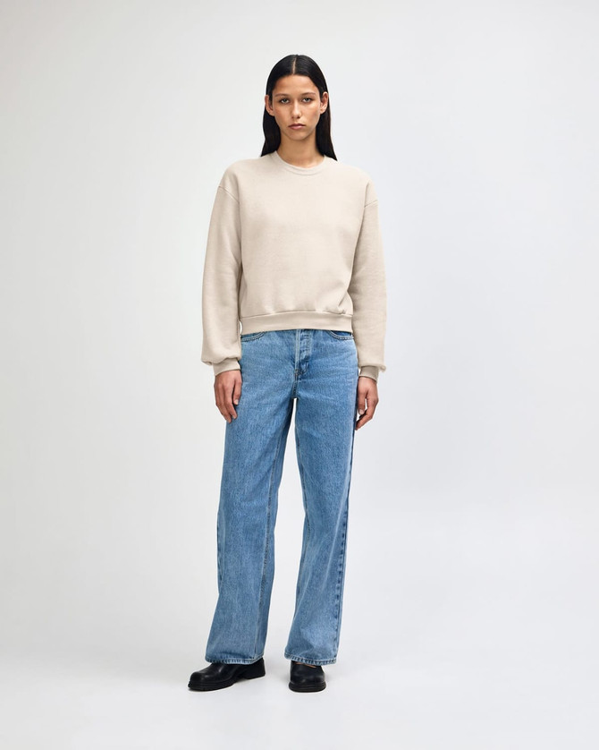 A women's crewneck sweatshirt in a light beige colour, styled with blue jeans and white shoes. It has a small logo.