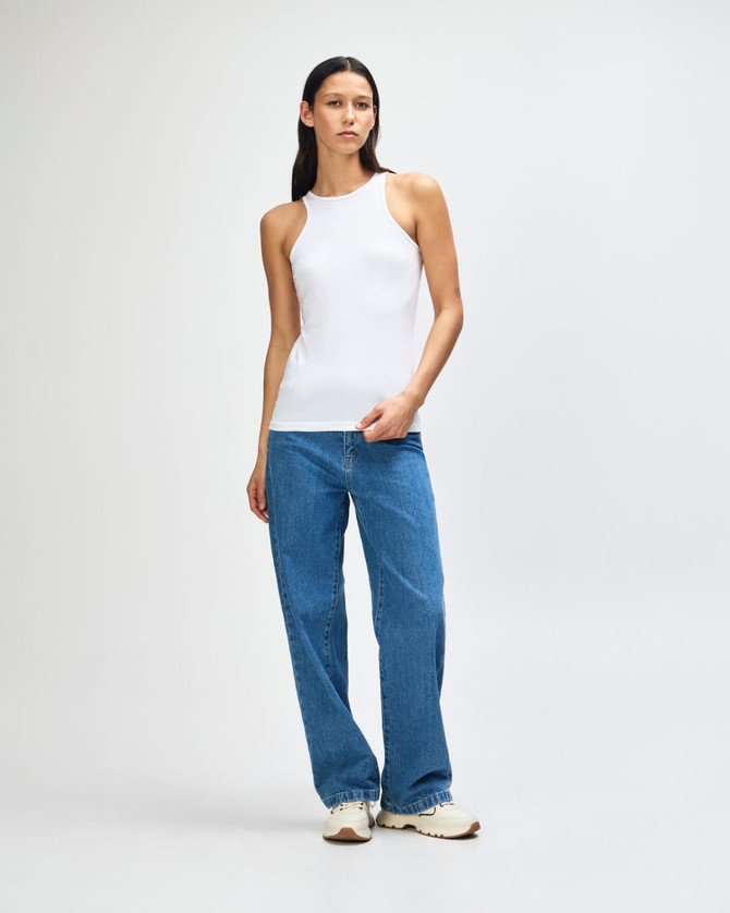 A white women's racerneck tank top worn with light blue wide-leg jeans, featuring a logo on the tank.