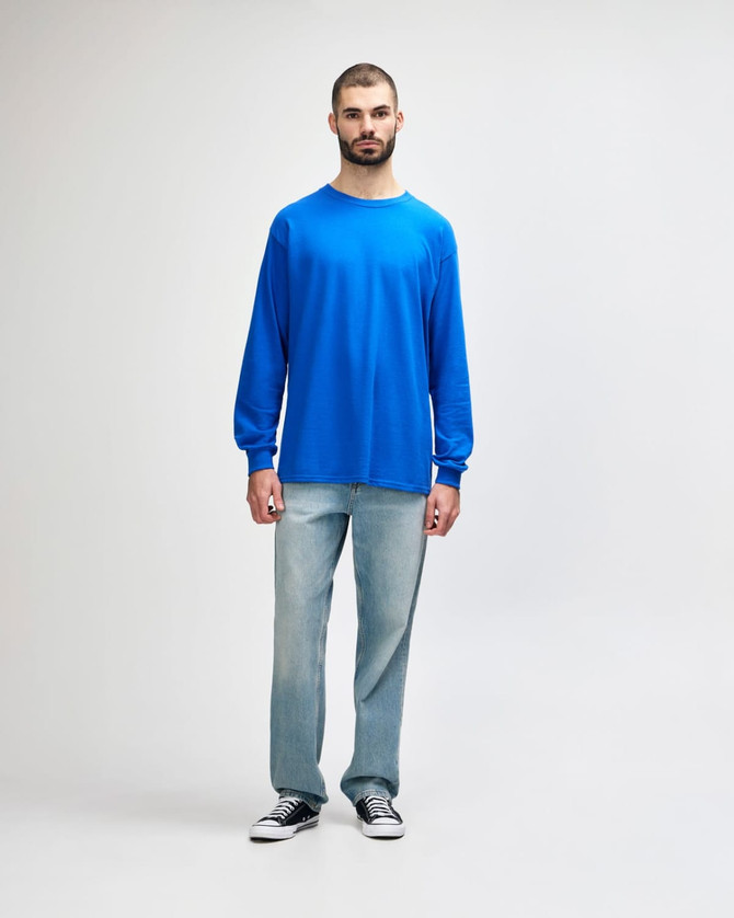 A man wearing a blue long sleeve T-shirt and light blue jeans, standing against a neutral background.