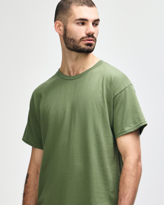 A man models a Gildan Heavy Cotton Adult T-Shirt in olive green, featuring a simple design and a logo.