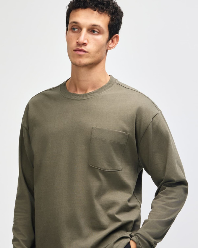 A model wearing an olive green American Apparel Super Heavyweight T-Shirt with a pocket. The shirt features long sleeves.