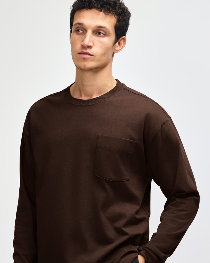 A model wears a brown American Apparel Super Heavyweight T-Shirt with a chest pocket. The image features a logo.