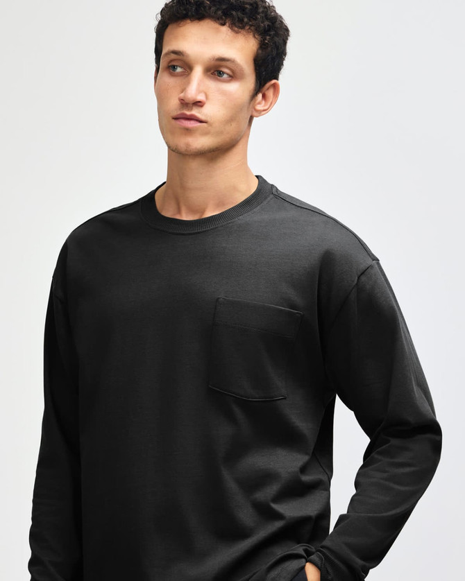 A black super heavyweight t-shirt featuring a pocket, modelled by a person with curly hair. It has a logo displayed.