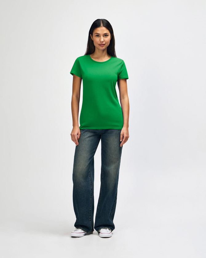A woman stands wearing a green Gildan Heavy Cotton Ladies' T-Shirt and blue jeans, with a casual pose and white shoes.