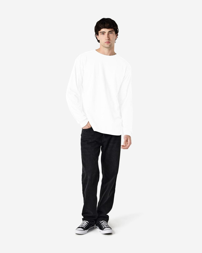 A model wearing a white long sleeve T-shirt with black jeans and sneakers, standing against a light background.