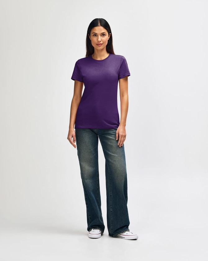 A woman is wearing a purple Gildan Softstyle ladies' T-shirt paired with blue jeans and white sneakers.