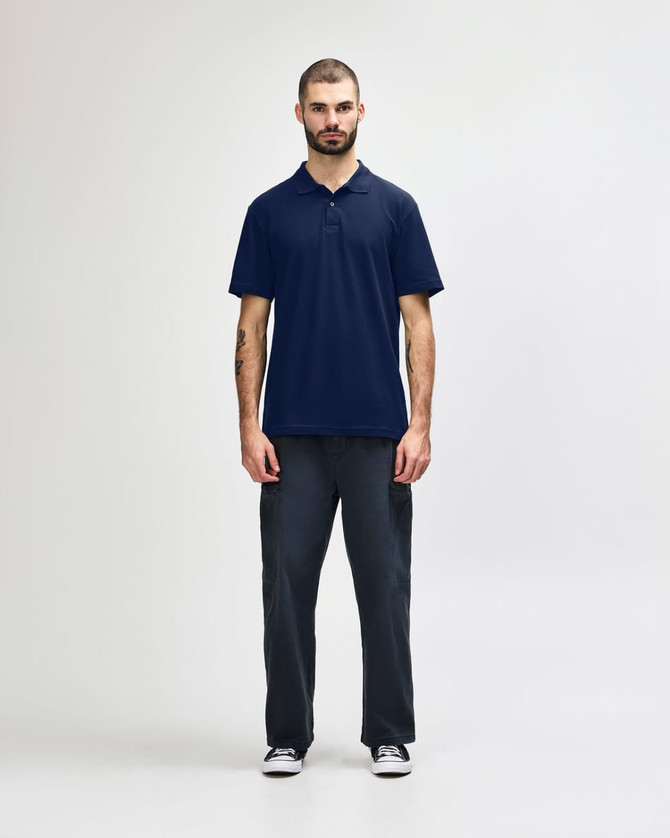A man stands wearing a navy blue Gildan Softstyle adult double pique polo and black jeans. The polo features a logo.