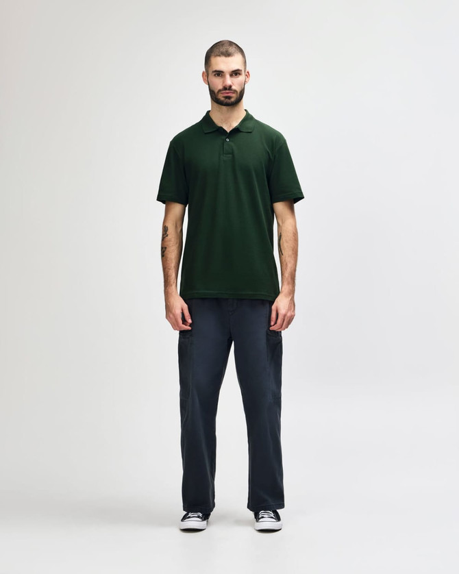 A man stands wearing a green Gildan Softstyle adult double pique polo, paired with black pants and shoes.