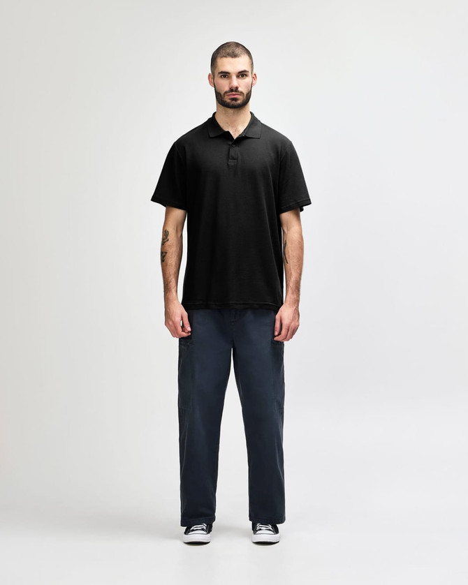 A model wearing a black Gildan Softstyle Adult Double Pique Polo, paired with dark jeans and sneakers.