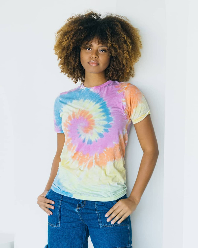 A tie-dye adult t-shirt in pastel colours, featuring a spiral design. A model poses while leaning against a wall.