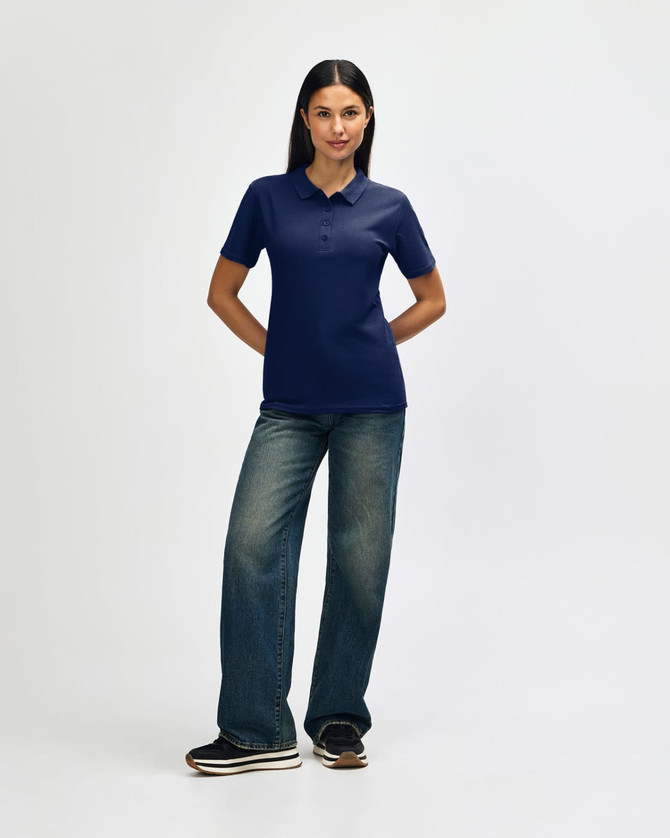 A woman stands smiling in a navy blue Gildan Softstyle ladies' polo shirt and denim jeans, wearing white sneakers.