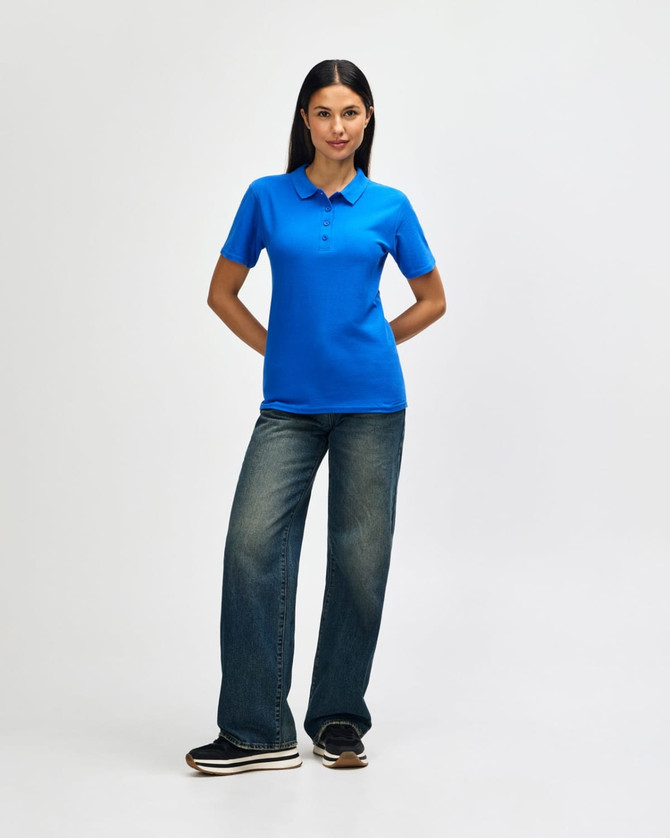 A woman poses in a bright blue polo shirt and jeans, standing confidently with arms crossed. The polo features a logo.
