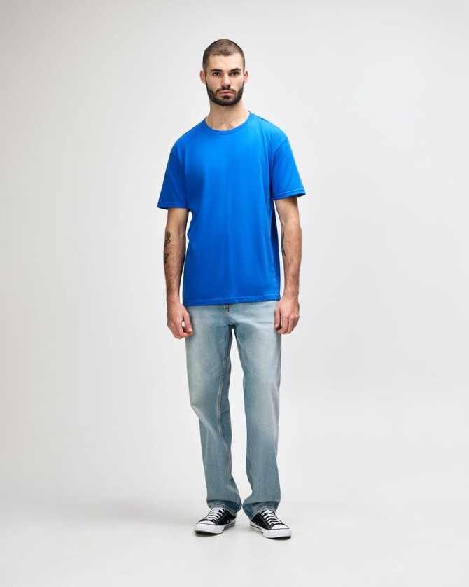 A man wearing a blue Gildan Softstyle adult midweight T-shirt paired with light denim jeans and white shoes.