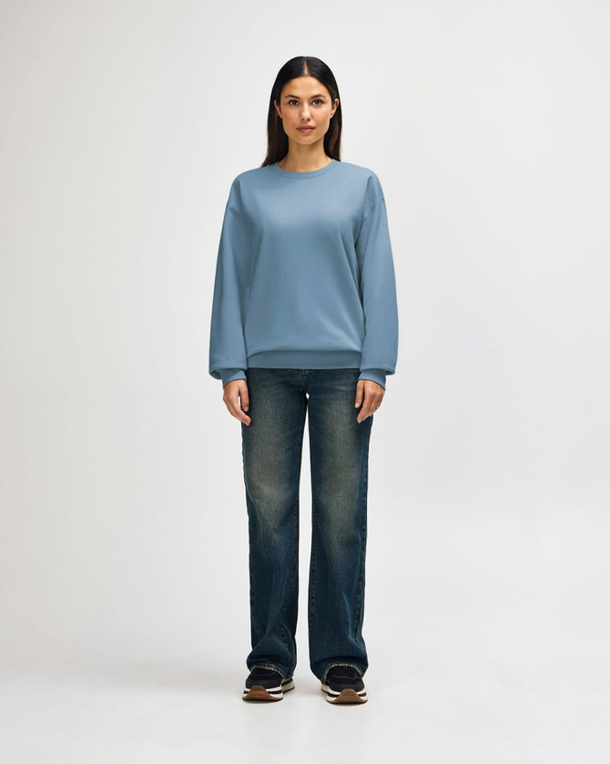 A woman stands wearing a light blue Gildan Softstyle sweatshirt paired with blue jeans and white sneakers.