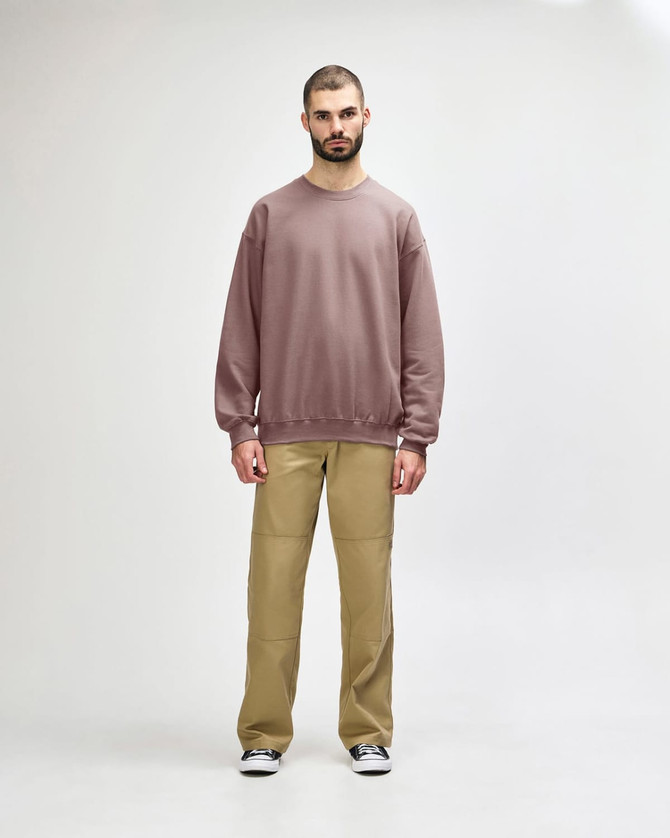 A male model wears a dusty pink Gildan Softstyle Adult Sweatshirt with black pants and black sneakers. The sweatshirt has a simple design.