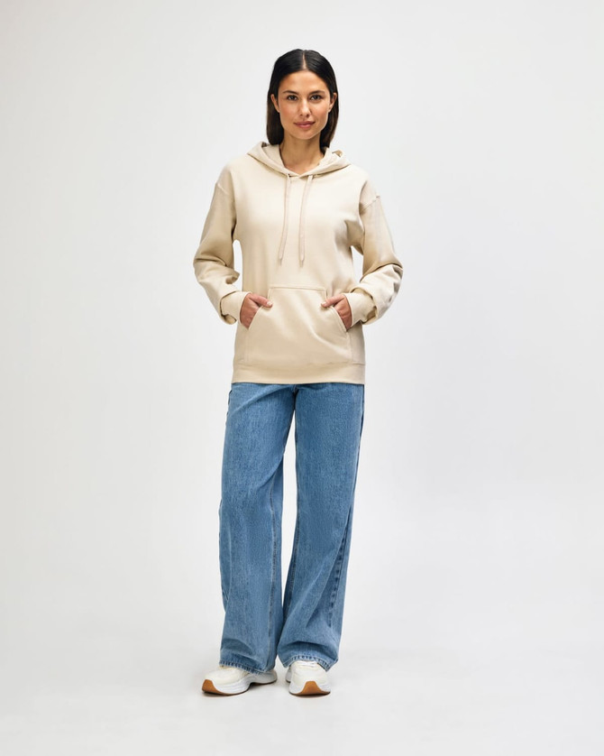 A hooded sweatshirt in cream worn by a person, paired with blue jeans and white shoes. The sweatshirt has a logo.