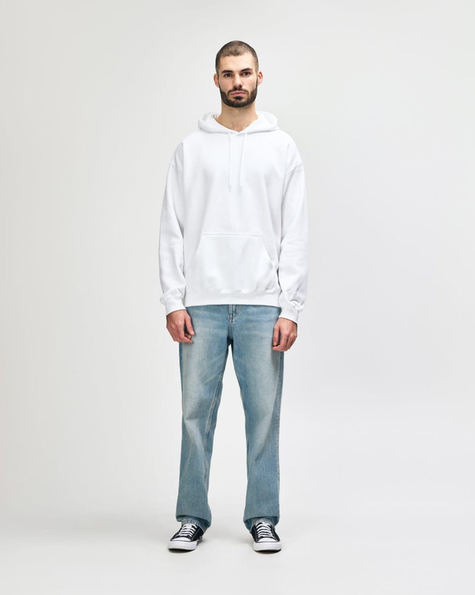A man stands in a white hooded sweatshirt and light blue jeans, wearing black shoes. The sweatshirt has a logo.