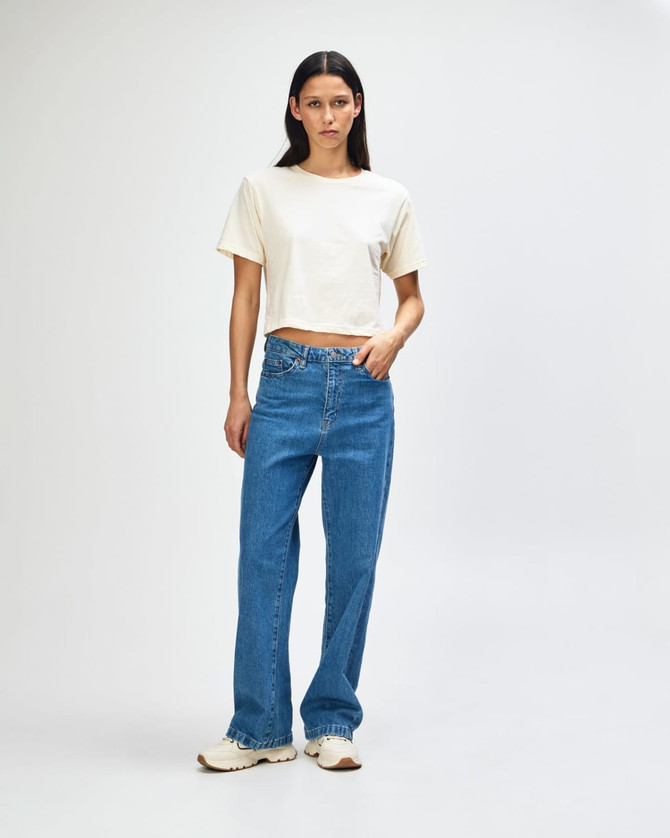A woman stands in a beige boxy t-shirt and light blue cargo jeans, featuring a logo on the shirt.
