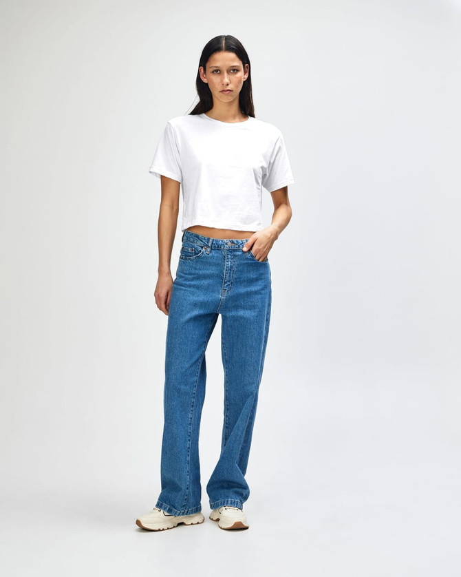 A woman stands wearing a white boxy t-shirt and light blue wide-leg jeans, featuring a logo on the attire.