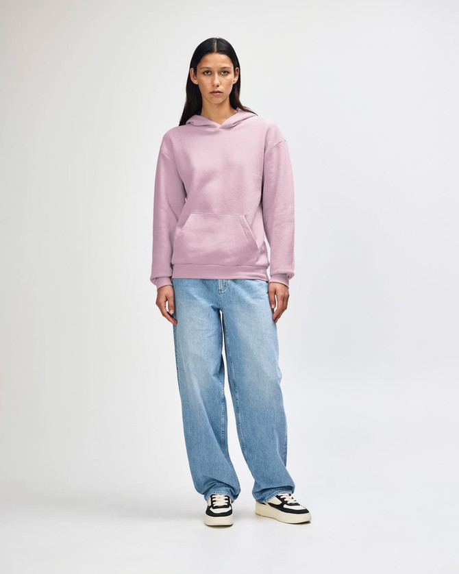 A unisex hooded sweatshirt in pink, featuring a front pocket and a logo, worn with light blue jeans.
