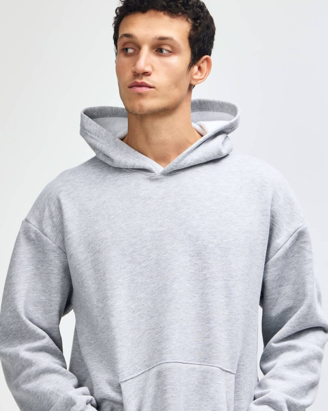 A grey unisex hooded sweatshirt with a front pocket and a logo. The model is standing with hands in pockets.