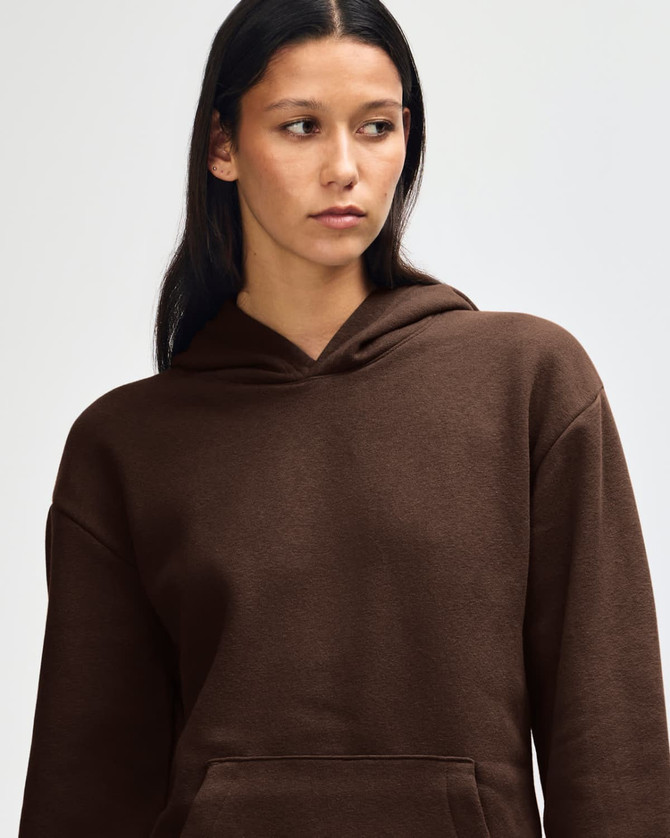 A brown unisex hooded sweatshirt with a front pocket and a model adjusting the hood. It features a logo.